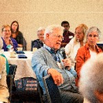 A man speaks into a microphone while a dozen people are seated around him listening to his question